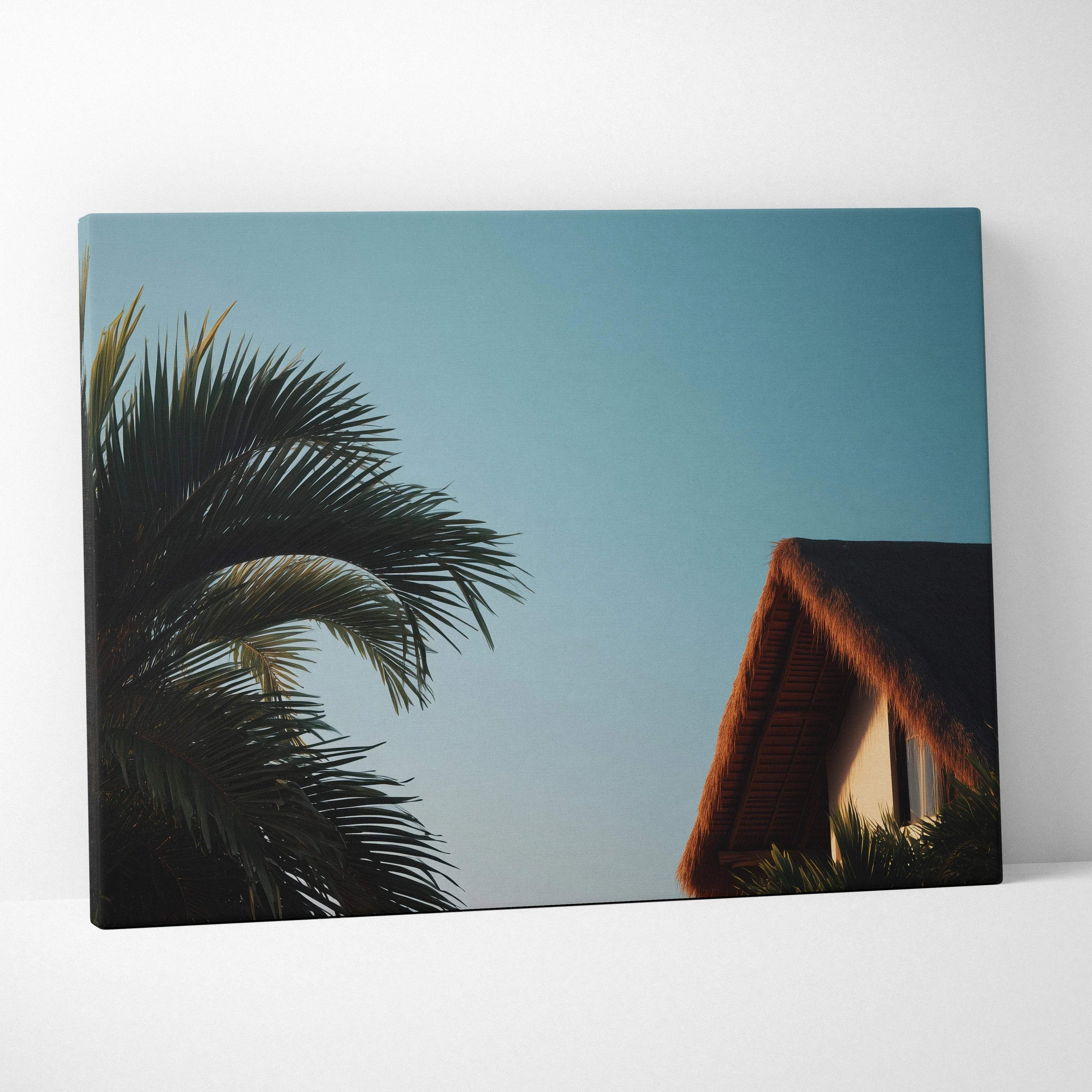 Canvas wall art featuring dark green palm tree leaves and a brown thatched roof house against a clear blue sky.