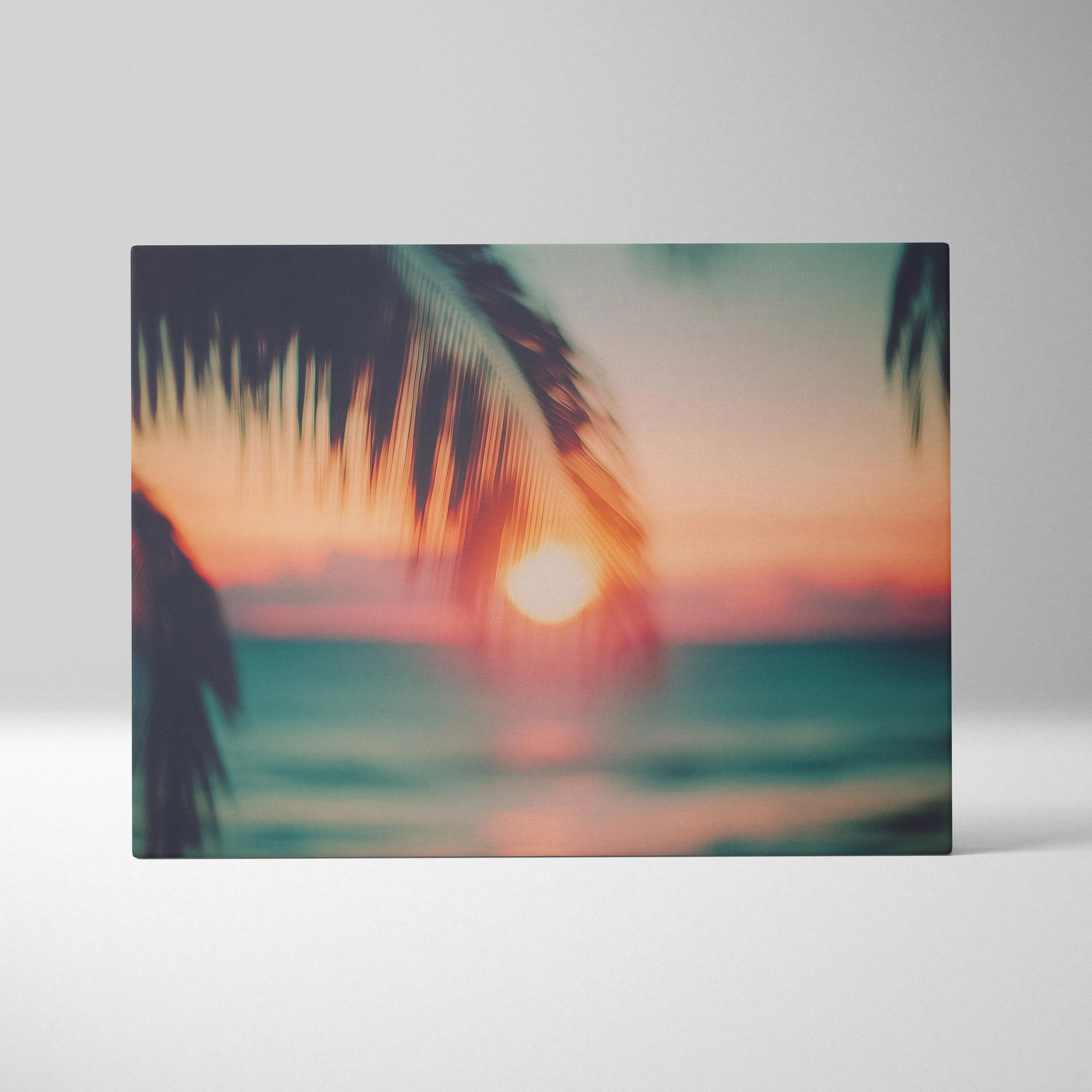 Canvas wall art featuring a blurred tropical sunset with warm orange, pink, and teal hues framed by silhouette palm leaves.