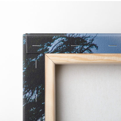 Light-colored wooden frame with canvas print of dark tree silhouettes against blue sky green blue mountain lake decor