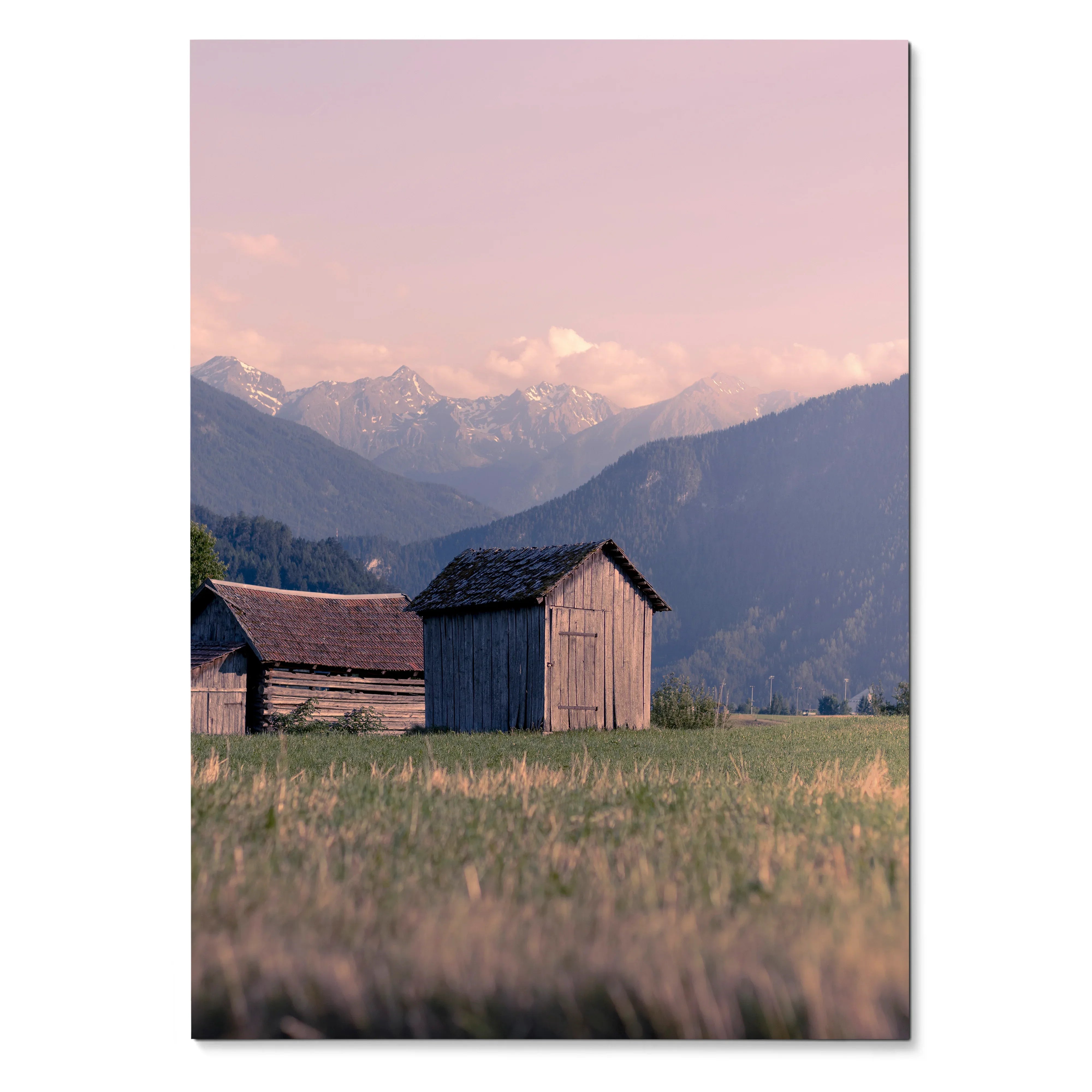 Vertical poster wall art of rustic wooden barns in a grassy field with mountain range and soft pink sky background.