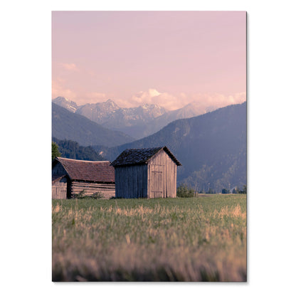 Vertical poster wall art of rustic wooden barns in a grassy field with mountain range and soft pink sky background.