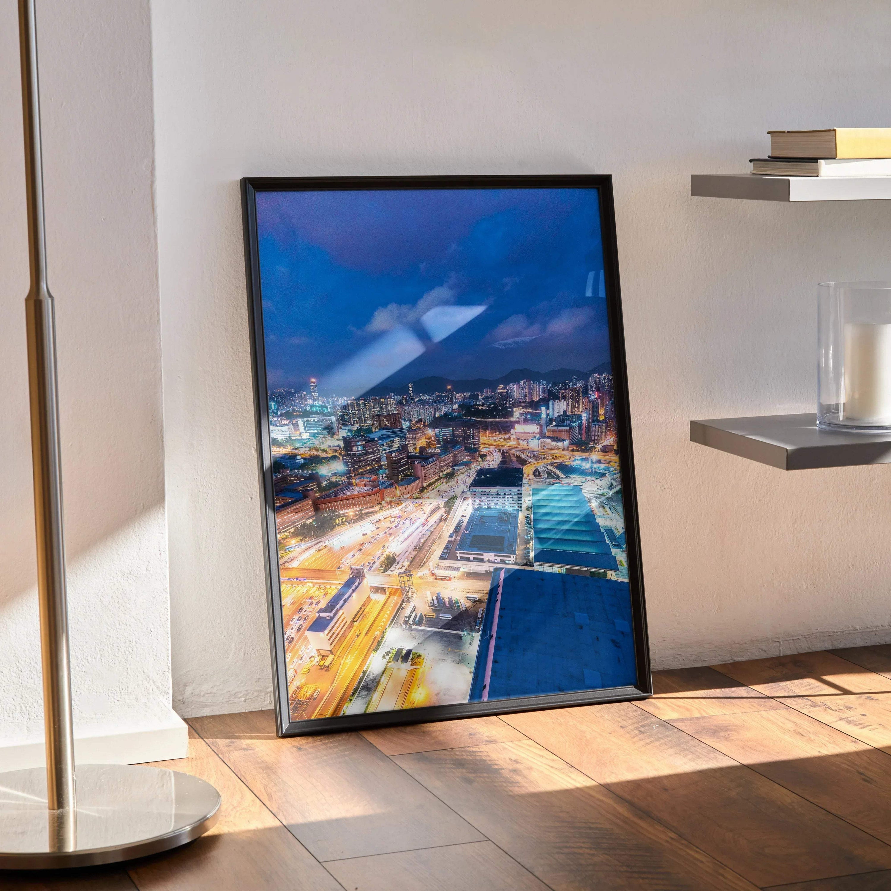 Urban night skyline wall art poster in black frame resting on hardwood floor by wall and modern shelves.