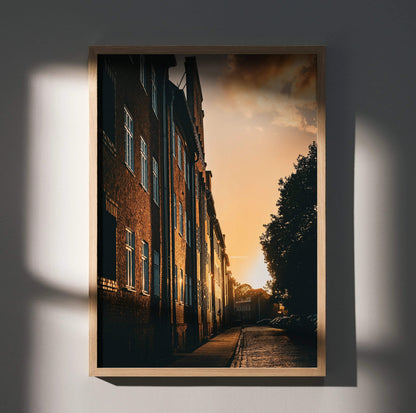 Vertical wall art poster of a sunset cityscape with warm light on brick buildings and trees.