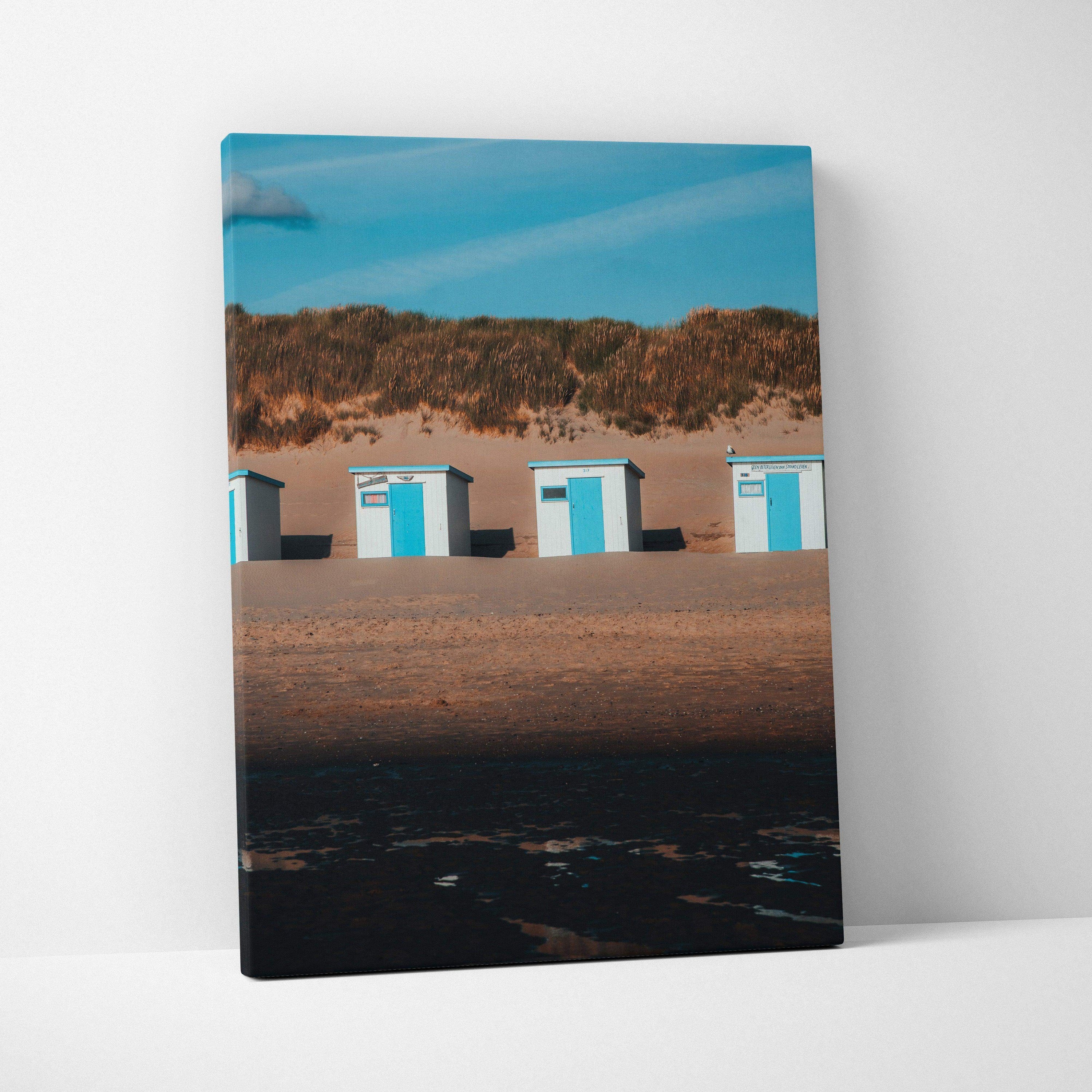 Vertical beach scene canvas wall art featuring blue and white beach huts against sand dunes and clear sky.