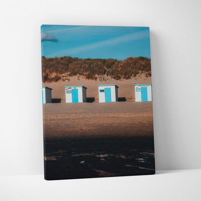 Vertical beach scene canvas wall art featuring blue and white beach huts against sand dunes and clear sky.