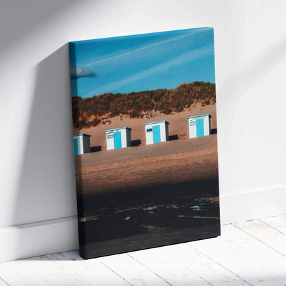 Vertical canvas wall art featuring white and blue beach huts against sand dunes under a clear blue sky.