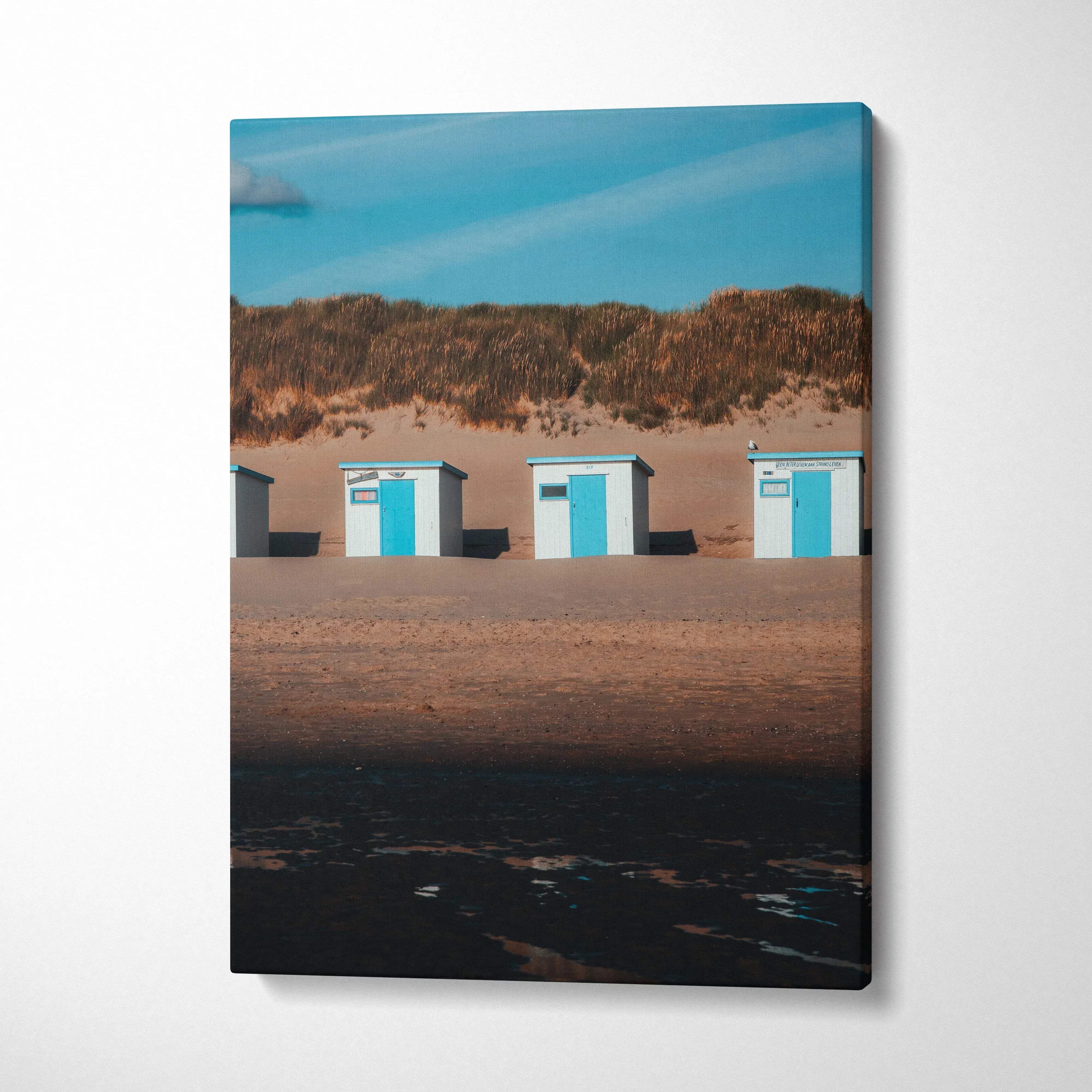 Vertical beach scene canvas wall art with blue sky, sand dunes, and three white and blue beach huts.