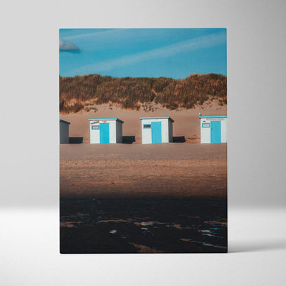 Vertical canvas wall art featuring three blue and white beach huts against sandy dunes and clear blue sky.