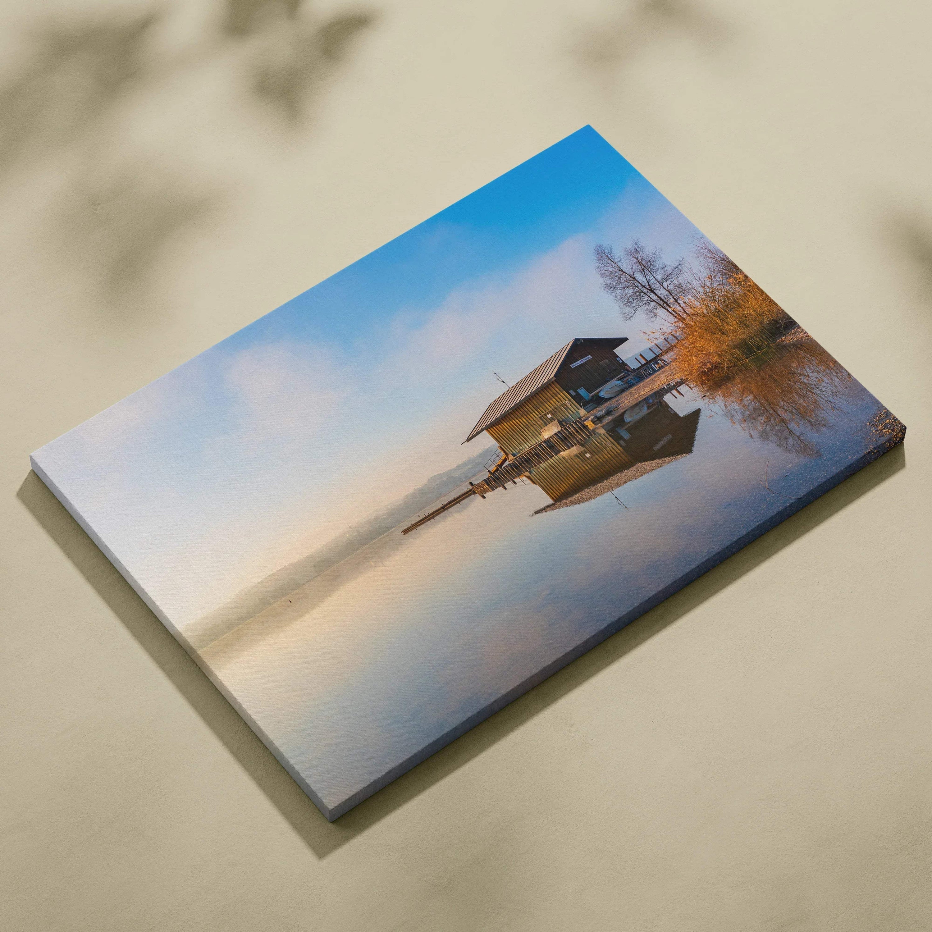 Canvas wall art featuring a serene lakeside cabin reflected in calm water under a soft blue sky.