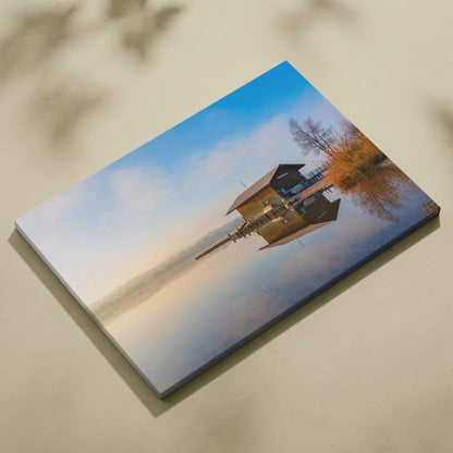 Canvas wall art featuring a serene lakeside cabin reflected in calm water under a soft blue sky.