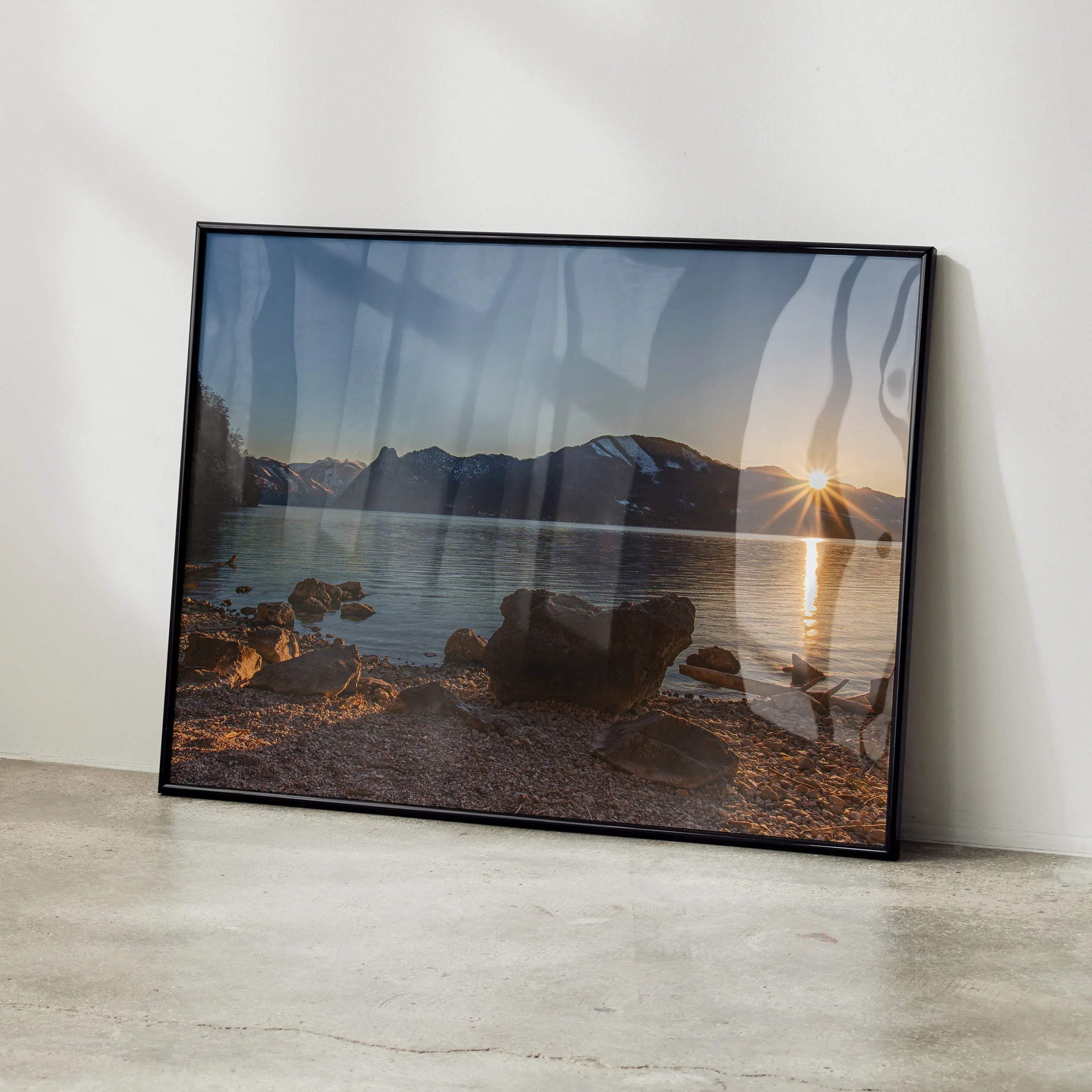 Framed wall art poster featuring a mountain sunset over a rocky lake shore with sun rays and calm water reflection.