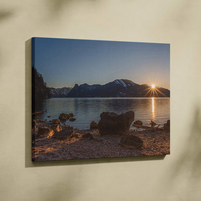 Canvas wall art of mountain sunset over calm lake with rocky shoreline and sunburst light effect.